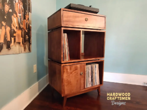 Tall solid maple record cabinet with upper turntable shelf, dovetail drawer, four divided storage compartments, one lower hardwood door, and tapered mid-century legs.