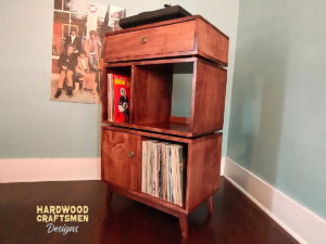 Solid cherry record storage cabinet with dovetail drawers, with upper turntable shelf, dovetail drawer, four divided storage compartments, one lower hardwood door, and tapered mid-century legs.