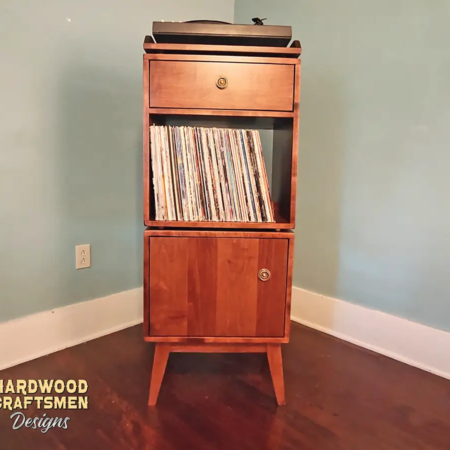 Tall, narrow solid cherry record player stand featuring a custom upper turntable shelf. The lower section includes a drawer above two storage compartments stacked vertically with a door on the bottom compartment, supported by tapered mid-century legs.