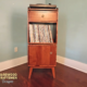Tall, narrow solid cherry record player stand featuring a custom upper turntable shelf. The lower section includes a drawer above two storage compartments stacked vertically with a door on the bottom compartment, supported by tapered mid-century legs.