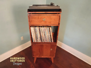 Narrow solid cherry record stand with upper custom turntable shelf, dovetail drawer, two divided storage compartments, one lower hardwood door, and tapered mid-century legs.