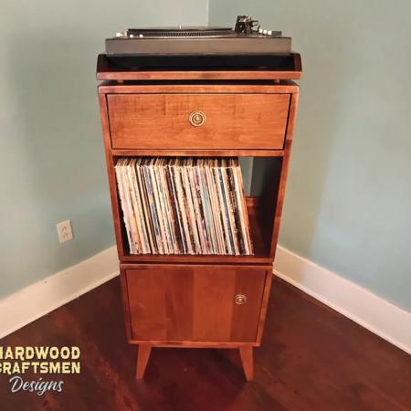 Narrow solid cherry record stand with upper custom turntable shelf, dovetail drawer, two divided storage compartments, one lower hardwood door, and tapered mid-century legs.