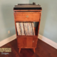 Narrow solid cherry record stand with upper custom turntable shelf, dovetail drawer, two divided storage compartments, one lower hardwood door, and tapered mid-century legs.