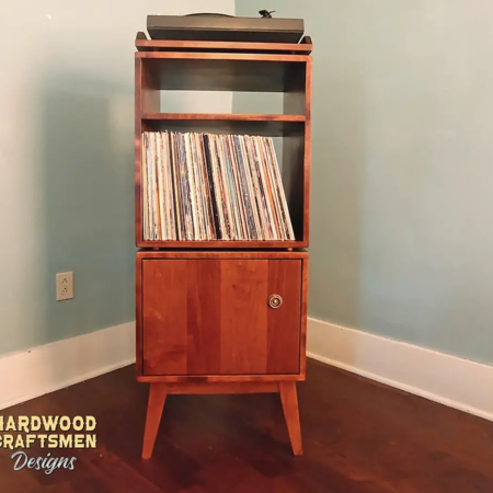 Tall, narrow solid cherry media console featuring a custom upper turntable shelf. The lower section includes a media compartment above two storage compartments stacked vertically with a door on the bottom compartment, supported by tapered mid-century legs.