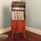 Tall, narrow solid cherry media console featuring a custom upper turntable shelf. The lower section includes a media compartment above two storage compartments stacked vertically with a door on the bottom compartment, supported by tapered mid-century legs.