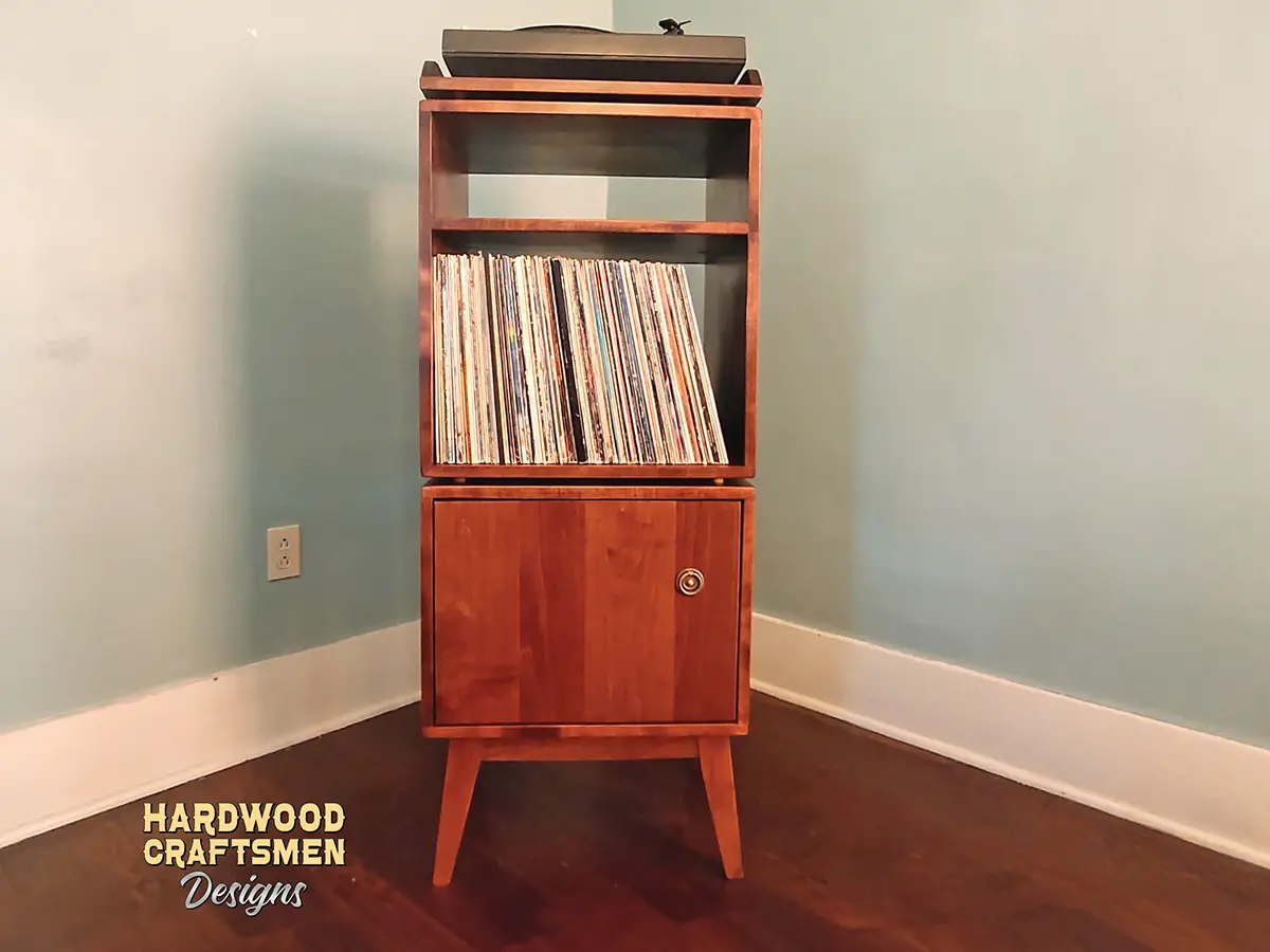 Tall, narrow solid cherry media console featuring a custom upper turntable shelf. The lower section includes a media compartment above two storage compartments stacked vertically with a door on the bottom compartment, supported by tapered mid-century legs.