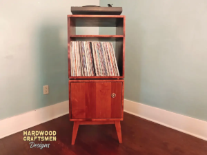 Narrow solid maple record stand with upper turntable shelf, dovetail drawer, two divided storage compartments, one lower hardwood door, and tapered mid-century legs.