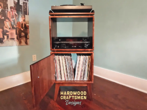 Tall, narrow solid maple record cabinet featuring a custom upper turntable and media equipment shelf below. The lower section includes two divided storage compartments stacked vertically with a door on the bottom compartment, all supported by tapered mid-century legs.