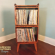Tall, narrow solid maple record cabinet featuring a custom upper turntable shelf. The lower section includes two divided storage compartments stacked vertically, supported by tapered mid-century legs.