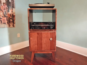 Tall, narrow maple record cabinet with an upper turntable shelf, a lower equipment shelf, and a single-door bottom cabinet enclosing two stacked storage compartments, supported by tapered mid-century legs.