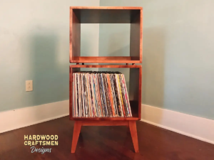 Tall, narrow solid maple record cabinet featuring an upper turntable and media equipment shelf. The lower section includes two open divided storage compartments stacked vertically, supported by tapered mid-century legs.