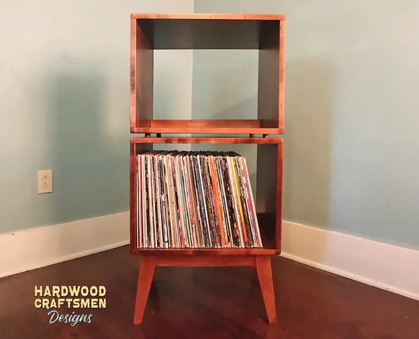 Tall, narrow solid maple record cabinet featuring an upper turntable and media equipment shelf. The lower section includes two open divided storage compartments stacked vertically, supported by tapered mid-century legs.