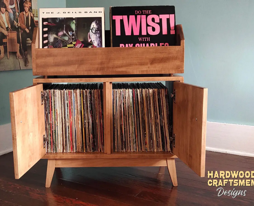 Large light-stained solid maple record cabinet featuring two upper vinyl flip bins. The lower section shows two open doors revealing two divided storage compartments, all supported by tapered mid-century legs.