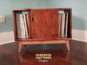 Low-profile solid maple record cabinet with a turntable shelf, 2 stacked storage bins, 2 sliding doors, and tapered wooden legs.