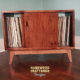 Low-profile solid maple record cabinet with a turntable shelf, 2 stacked storage bins, 2 sliding doors, and tapered wooden legs.