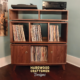 Solid walnut vinyl record storage cabinet featuring two upper side-by-side turntable and media compartments. The lower section includes six divided storage compartments with two outer doors on the bottom level, all supported by tapered mid-century legs.