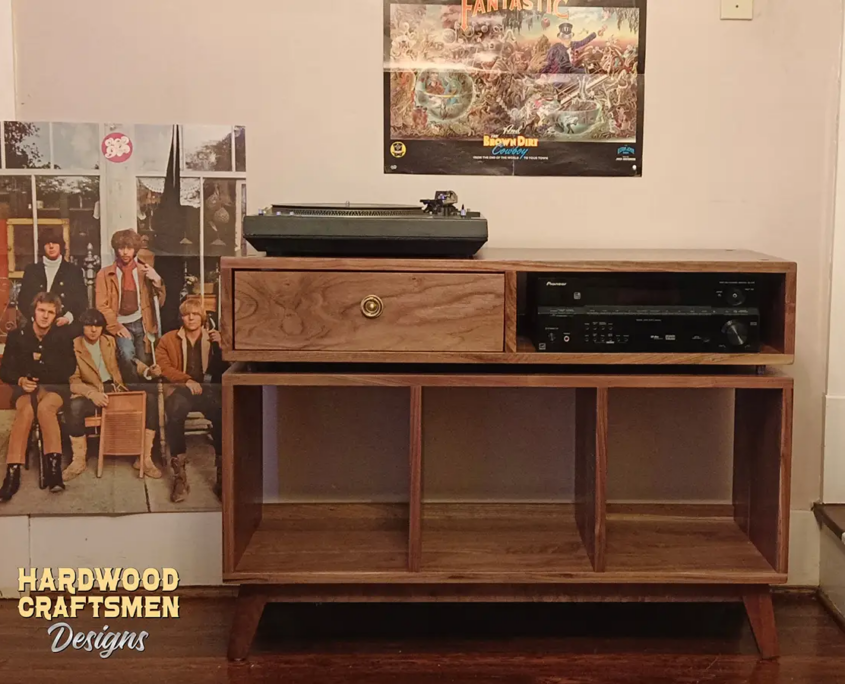 Large solid walnut media console, available from 42 to 72 inches, featuring an upper TV shelf. Below is a drawer and adjacent media compartment above three open storage compartments revealing the interior walls, all on tapered mid-century legs.