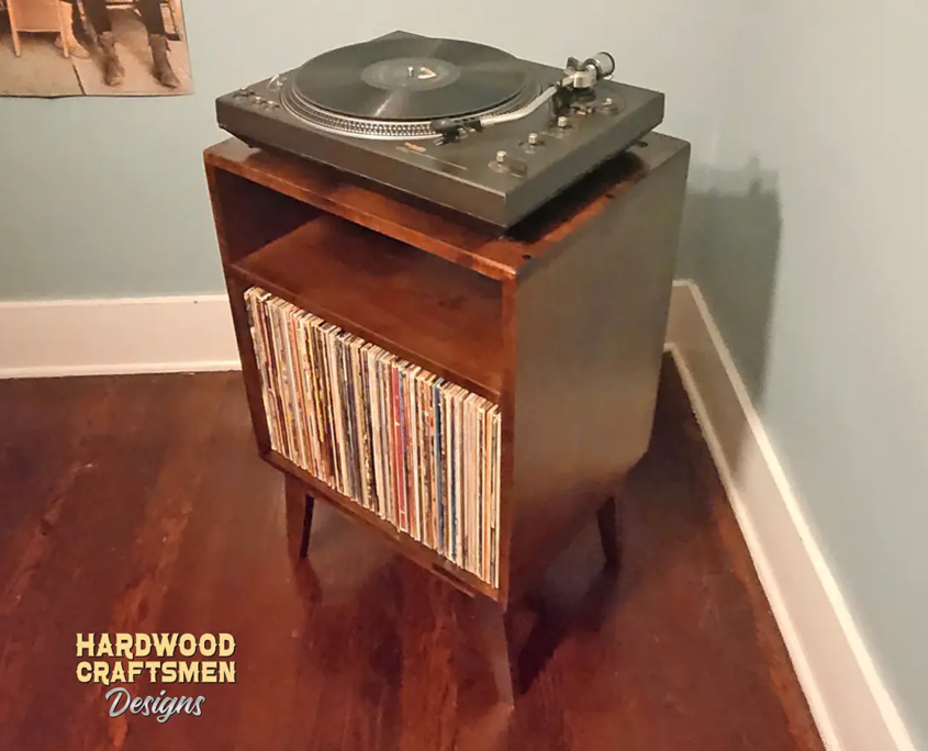 An angled view of a small, narrow solid cherry dark-stain media console featuring an upper turntable shelf. The lower section includes an open media cabinet with a storage compartment below, all supported by tapered mid-century legs.