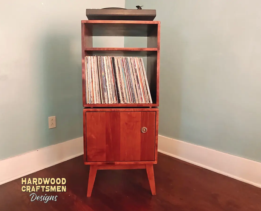 Tall, narrow solid cherry media console featuring an upper turntable shelf. The lower section includes a media compartment above two storage compartments stacked vertically with a door on the bottom compartment, supported by tapered mid-century legs.