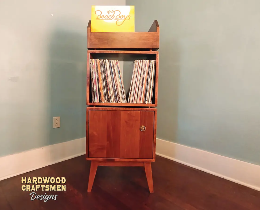 Narrow solid maple record cabinet featuring an upper vinyl flip bin. The lower section includes two vertically stacked storage compartments, with a closed door covering the bottom compartment, all supported by tapered mid-century legs.