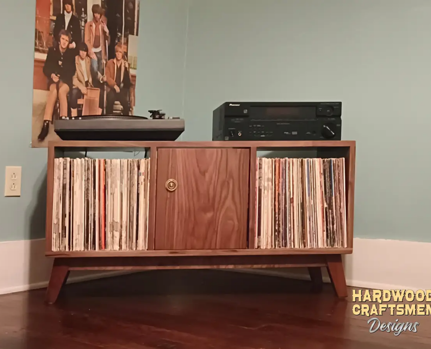 Low-profile solid walnut media console, available from 42 to 72 inches, featuring an upper TV shelf above three divided storage compartments, with one center hardwood door covering the center compartment, all on tapered mid-century legs.