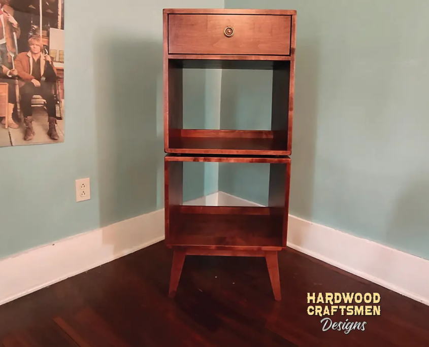 Tall, narrow solid cherry record cabinet featuring an upper turntable shelf. The lower section includes a drawer above two open storage compartments stacked vertically, supported by tapered mid-century legs.