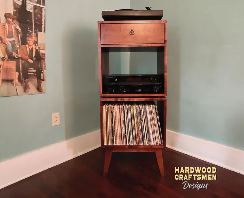 Tall, narrow solid cherry record cabinet featuring an upper turntable shelf. The lower section includes a drawer above a media shelf and an open storage compartment stacked vertically, supported by tapered mid-century legs.