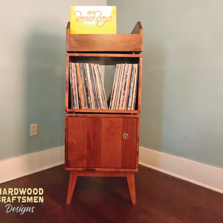Narrow solid maple record stand with upper vinyl record flip bin, two modular storage compartments, one lower hardwood door, and tapered mid-century legs.